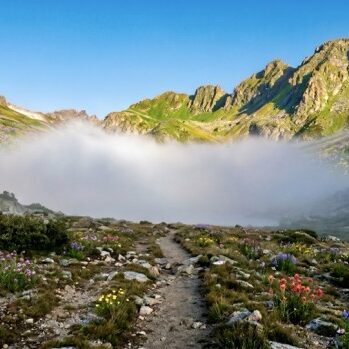 Mountain Trail Being Safe Blog Post Featured Image A trail through a mountain pass with fog ahead and clear sky behind — the cost of sitting in cash and finding the path forward, Riverwater Partners