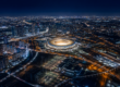 Aerial night view of a brightly lit stadium at the center of a sprawling, illuminated city grid