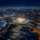 Aerial night view of a brightly lit stadium at the center of a sprawling, illuminated city grid